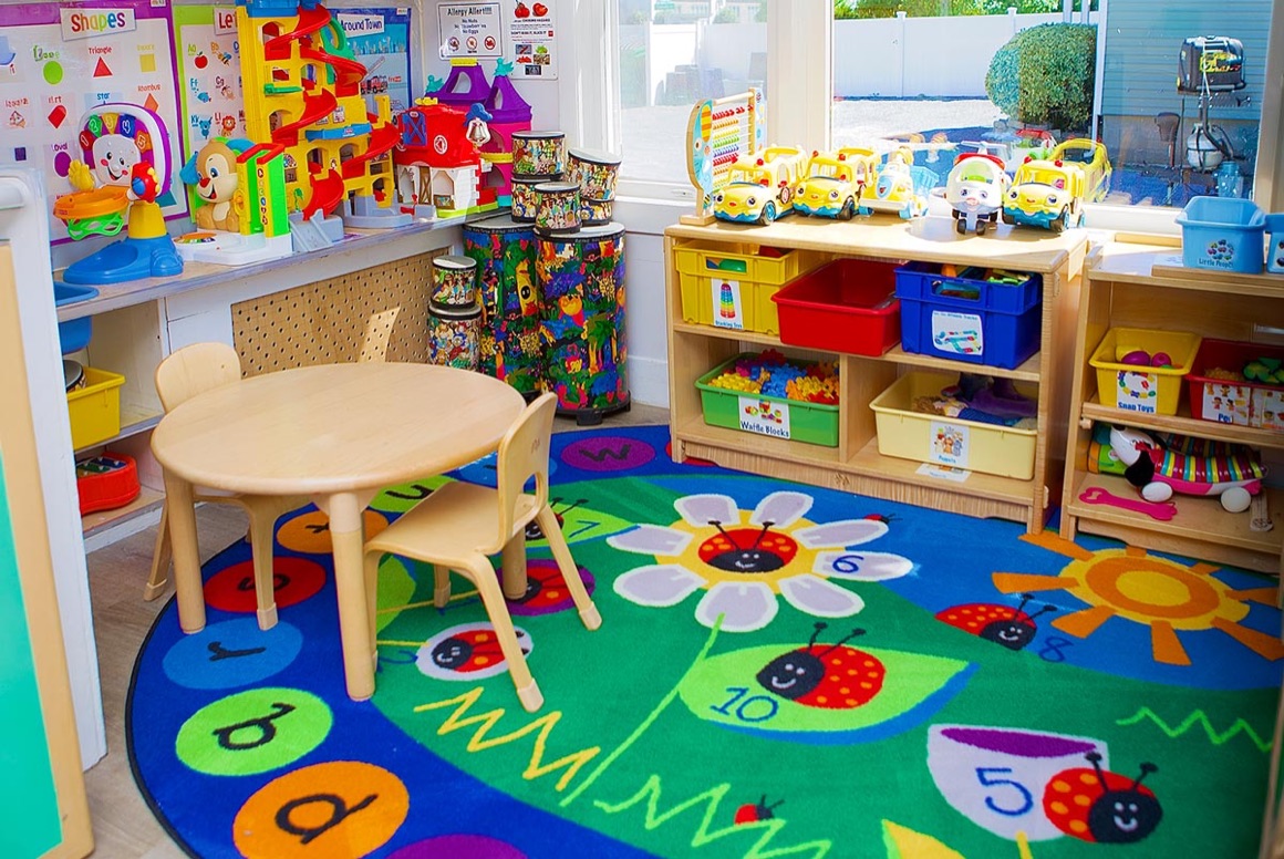 A classroom activity area with a number rug, toy shelves, and a small table by the window