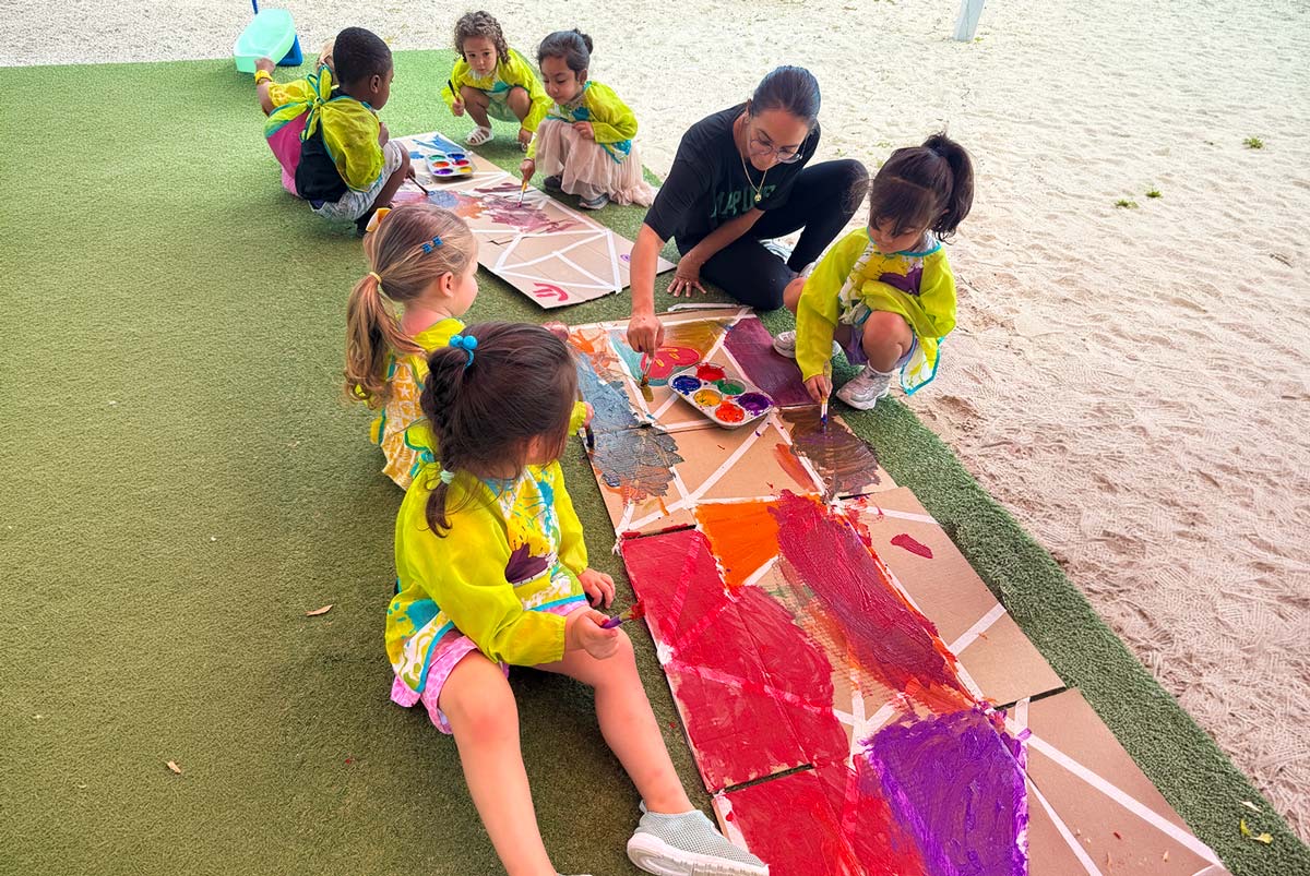 Preschoolers and a teacher painting a large cardboard kite outdoors