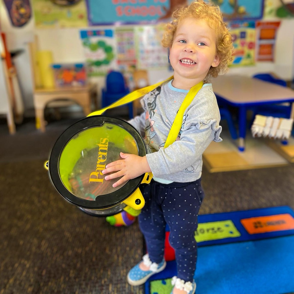 A toddler smiling while playing a drum during a music enrichment session